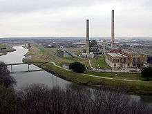 A middle-sized river flows by a factory with two tall smokestacks.