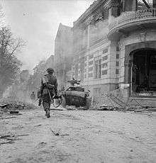 A man walks away from the camera, down a street littered with debris.  Ahead of him is a small armoured vehicle.
