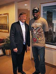 Donald Trump posing with basketball personality Dennis Rodman in a room with paintings adorning the walls. Trump is wearing a suit with a light-colored tie and dress shirt, while Rodman is wearing a brown t-shirt with a design on it, blue jeans, and a baseball cap that also has a design on it.