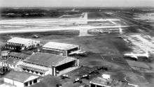Black-and-white photo of a runway and hangars. Only one large airplane can be seen.