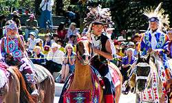 Colour photograph of Tsuu T'ina children in traditional costume on horseback at a Stampede Parade in front of an audience