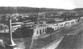 A grainy, black and white photograph showing several single story buildings next to a dirt road. Several homes and other buildings stretch toward the mountains in the distance. A chimney is seen in the foreground.