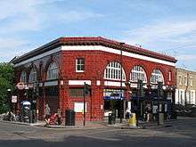 A two-storey station building in red glazed blocks sits at the corner of a road junction. Large semi-circular windows feature prominently on the upper floor.