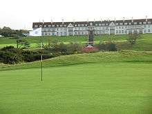 A wide, sprawling golf course. In the background is the Turnberry Hotel, a two-story hotel with white façade and a red roof. This picture was taken in Ayrshire, Scotland.