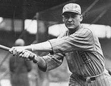 A man in a light-colored pinstriped baseball uniform looks into the camera having just swung his bat to the left.