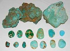 Three rough chunks of raw turquoise in brown matrix are at the top of the picture, below which are a range of thirteen finished cabochons showing various colors ranging from green to light turquoise blue, and a range of spiderweb matrix ranging from none to light yellow to deep brown.