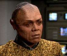The dark-skinned Tom Wright is in makeup and costume as Tuvix.  Black makeup is mottled across his cheeks and high brow, black straight hair is combed back from the hairline, pointed ear prosthetics have been applied, and amber-colored contact lenses are visible.  The costume is colored in mustard and black like Tuvok's, but patterned in flowers and cut to a style like Neelix's customary clothing.