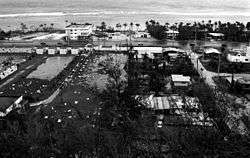 Elevated black and white photograph of businesses surrounding a shoreline road. Though the waves and sea can be seen in the background, floodwaters and strewn debris are visible in the foreground.