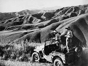 Two men in slouch hats standing in a jeep. They are staring at a series of rugged looking ridge lines