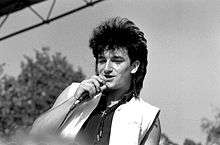 A black and white image of a light-skinned man singing into a microphone. He is visible from the chest up and wears a sleeveless black shirt with an opened sleeveless white vest overtop. A small cross is worn around his neck. His black hair is styled into a mullet. The man looks past the camera to the left. A mixture of trees and sky are visible in the background.