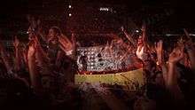 A close-up shot of fans inside a stadium during a concert, many of whom have their arms in the air. A transparent layer on top shows a concert stage with a curved video screen showing black-and-white images of musicians performing, with solid red and yellow colors below.