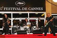 The Edge, Adam Clayton, and Larry Mullen, Jr. performing outside a building on red carpet, with a sign in the background reading "Festival de Cannes".