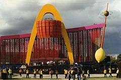 A concert stage in the daytime, with a pink video screen, tall golden arch, oversized lemon, and a stick with an olive through it.