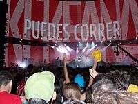 The view of the stage from the audience's perspective, surrounded by fans, with the words "PUEDES CORRER" visible on the video screen. A camera on the right side shows the concert being filmed.