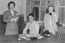 A young man seated flanked by two women standing, all of them behind a desk. The woman on the right is holding a telephone; the one on the left is holding what appears to be correspondence.