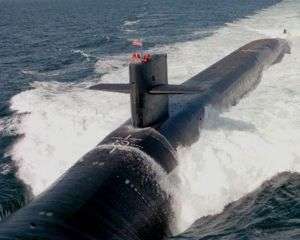 Submarine running at high speed of surface, creating huge waves. Several people's heads pop up from the boat's sail.