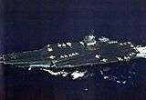 Large aircraft carrier with boxy island superstructure and numerous aircraft on its flight deck.