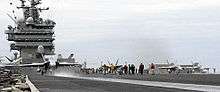 Three gray F/A-18 Hornet strike fighter aircraft line up across the frame for catapult launches from an aircraft carrier's deck. Support staff is seen on the deck throughout, while exhaust can be seen from the engines of the right-hand aircraft.