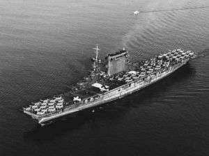 Left front oblique aerial view of a dirty grey aircraft carrier. Her full-length flight deck is covered by aircraft and she has an enormous funnel separate from her island.