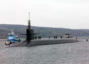 Surfaced submarine in bay with mountains in the background. Flanked to its starboard side by a tug, the boat has several people standing atop of it.