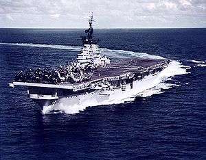 A color photo of a large aircraft carrier moving through the ocean with a deck full of aircraft