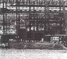 An incomplete submarine lays a rest; tall steel gantries and a dock dominate the background.