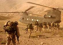A line of soldiers carrying equipment on their backs walking toward a transport helicopter in desert terrain