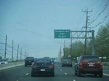A six lane divided highway lined with power lines. A green sign over the road reads Scudders Mill Road ¼ mile.