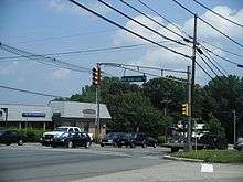 A four lane undivided road lined with businesses at a traffic light. A sign on the traffic light pole reads Old Budd Lake Road.