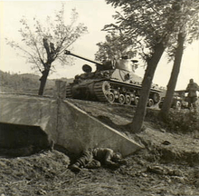A tank sits side-on on a raised road between two trees, followed by a smaller vehicle, while a soldier stands. In the foreground the body of another soldier lies slumped near a culvert below the road
