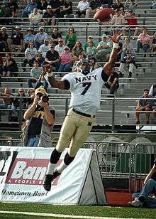 An American football player in a gold and white uniform strains to catch a football out of reach.
