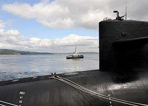 In the foreground: the aft part of the bow and forward half of the sail of a black submarine. In the middle distance, USS Reliant motoring toward the left side of the image.
