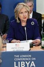 Hillary Clinton speaks at a London meeting to discuss NATO military intervention in Libya on March 29, 2011. She is standing behind a blue podium with a sign that has the words "THE LONDON CONFERENCE ON LIBYA" printed in white-on-blue text in capital letters.