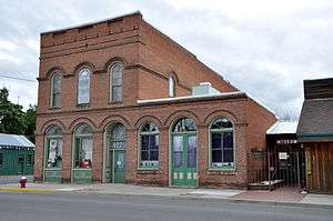 Photograph of a 2-story brick commercial building standing on a street corner, arched windows and doors