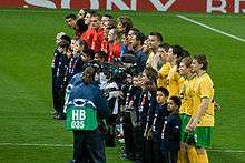 Celtic (foreground, in yellow) and Manchester United players (background, in red) and the match officials (in grey) line up prior to the group stage match between the two sides. Each player has a child mascot wearing a navy blue shirt standing in front of him, and a television camera crew is focusing on the Celtic players.