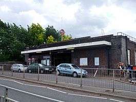 A brown-bricked building with a rectangular, dark blue sign reading "UPNEY STATION" in white letters all under a light blue sky with white clouds