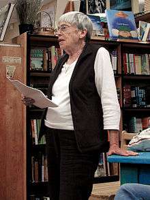 Photograph of Ursula K. Le Guin standing and reading aloud in a bookshop