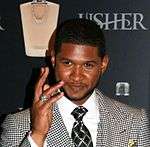 Usher, wearing a white shirt, criss-cross tie and chequered jacket, waving in front of his perfume branded wallpaper
