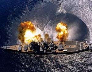 An overhead view of a large ship with a teardrop shape firing guns toward the top of the image.