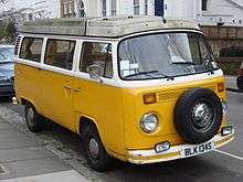 A yellow and white Volkswagen Type 2 vehicle with a dirty roof is parked on a street.