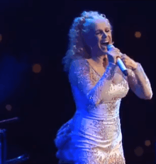 A woman with blond hair is wearing a white dress and is singing on a stage