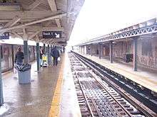 The platform level on a snowy day, on the center island platform. A train is stopped on the left-hand track. On the right is another, empty track, as well as a disused side platform with a white windscreen.