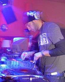 A DJ wearing headphones is performing using his turntables and other electronic equipment. A computer monitor is visible in the background.