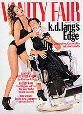 Cover of Vanity Fair from 1993 showing short-haired k.d. lang sitting in a barber chair with shaving foam on her chin, reclining wearing a pinstripe pair of pants with cuffs tucked into black lace boots, open-collar dress shirt, and black and white striped tie, holding a compact mirror. Her eyes are closed. Supermodel Cindy Crawford wears a one-piece black bathing suit and high heel boots, holds lang's face to her breast and has her head thrown back so her long hair cascades down her back. Crawford holds a straight razor to lang's chin.
