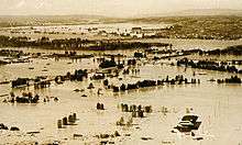 Floodwaters inundate a populated area. A few roadways and partly submerged buildings are visible in the midst of the flood. A long bridge crosses a flooding river in the distance. Beyond the river the ground rises, and buildings there appear to be above water.