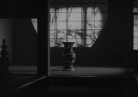 In a semi-darkened room, an ordinary vase is seen on the floor in the background, behind which is a screen upon which the shadows of branches appear