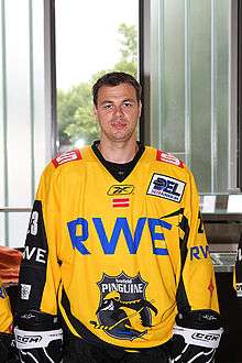 An ice hockey player standing directly in front of the camera, He has brown hair. He is wearing a yellow and black uniform with a large blue RWE and a black and yellow penguin on his chest.