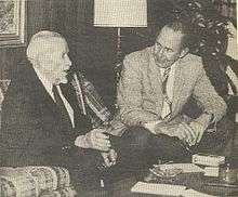 A black-and-white photograph of two men sitting by a low table talking to each other. The man on the left is much older, has white hair, and is wearing a dark suit with a white shirt and a dark tie. He sitting on a plaid couch and gesturing with his right hand as he speaks. The man on the left is younger, has dark hair, and is wearing a light jacket, dark pants, a white shirt and a patterned tie. He is sitting on a chair with his arms resting on his legs as he leans forward to listen to the other man.