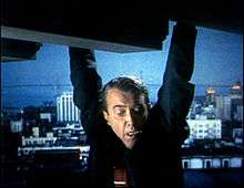 A man, seen from mid-chest up, hangs by his hands from the edge of an apparently tall structure, gazing down in fear. He is wearing a dark suit and an orange tie with a clip. In the distance behind him is a cityscape at night or in the early morning. There is a bluish cast to the background.