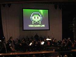 A photograph of an orchestra on a dimly lit stage. Above the group is a projection screen with a black, white, and green image of pixel art. The pixel art is an oval object wearing headphones with eyes and four tentacles. Below the pixel art is the phrase "Video Games Live".